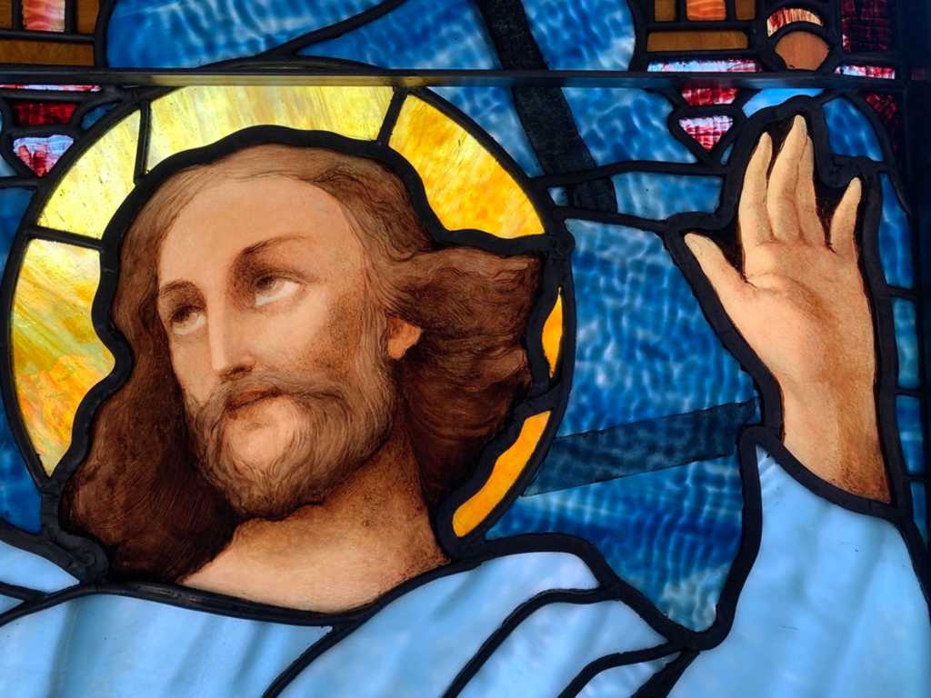 Stained glass window depicting Jesus Christ with a serene expression, raised hand, and a radiant halo, showcasing intricate artistry and vibrant colors, symbolizing spiritual themes in church worship.