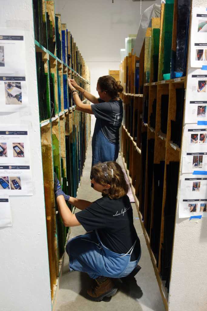 Artisans restoring stained glass windows in workshop, handling colored glass panels, surrounded by shelves of materials and reference images, emphasizing traditional craftsmanship and meticulous techniques.
