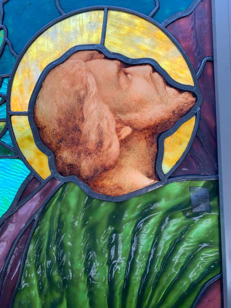 Stained glass window depicting a serene figure with flowing hair and green robes, showcasing intricate detailing and vibrant colors, reflecting craftsmanship in stained glass artistry.