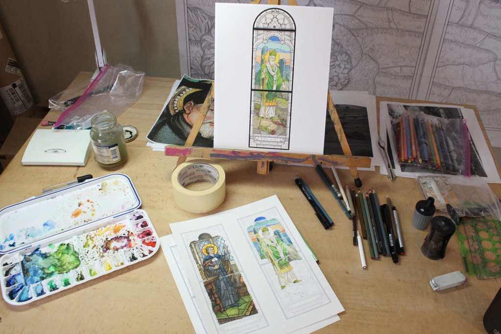 Artistic workspace with stained glass design sketches, watercolor palette, and drawing tools, showcasing the creative process at Willet Stained Glass Studios.