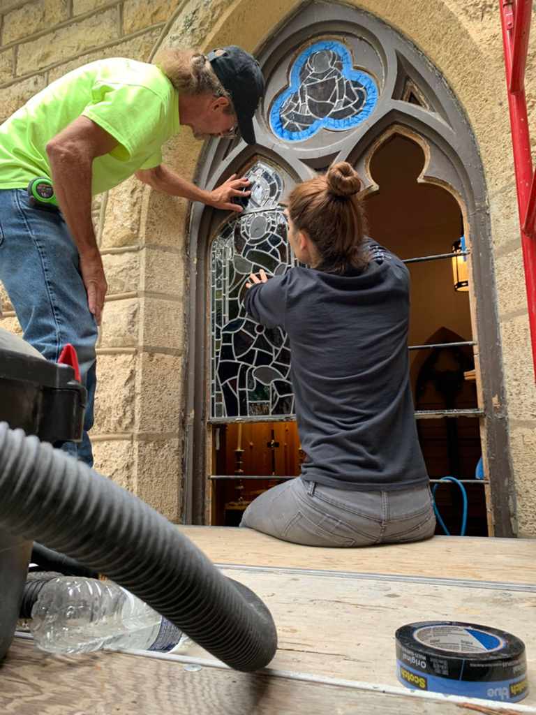 Technicians restoring a stained glass window, focusing on intricate details and structural integrity, with tools and materials visible in a church setting.
