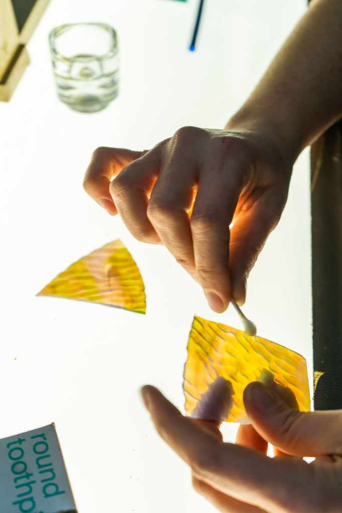 Hands cleaning stained glass pieces with a cotton swab, showcasing vibrant yellow glass and restoration tools on a work surface, emphasizing the meticulous repair process at Willet Stained Glass Studios.