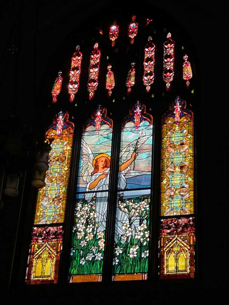 Stained glass window featuring an angel holding a flower, surrounded by floral designs and intricate patterns, showcasing the artistry of Willet Stained Glass Studios.