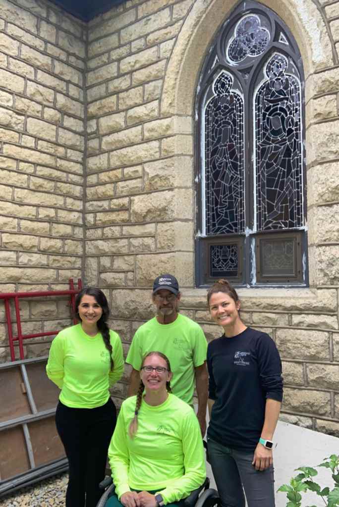 Team of stained glass restoration experts in bright shirts, standing in front of a historic stained glass window, showcasing craftsmanship and dedication to stained glass repair and preservation.