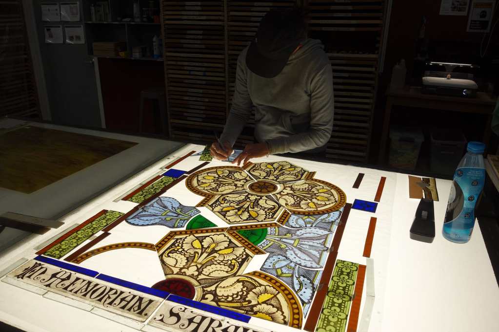 Artisan working on stained glass window design, showcasing intricate patterns and vibrant colors, with tools and materials arranged on a backlit work surface, emphasizing craftsmanship and restoration process at Willet Stained Glass Studios.
