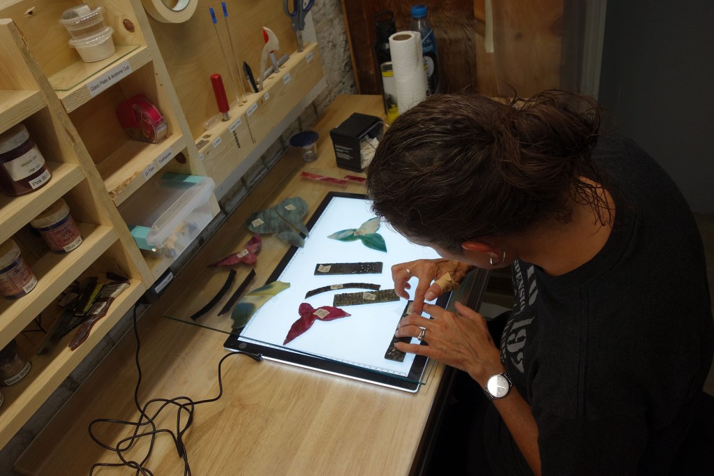 Woman working on stained glass pieces using a lightbox, surrounded by tools and glass materials, illustrating the craftsmanship and restoration process at Willet Stained Glass Studios.