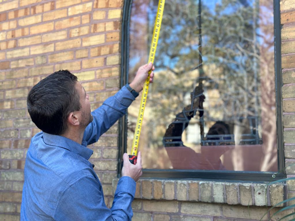 Man measuring a stained glass window for restoration, showcasing the intricate design and brick wall background, emphasizing the importance of stained glass maintenance.