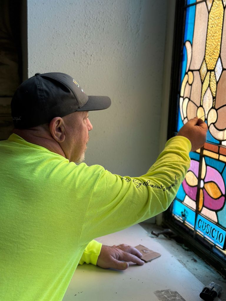 Restoration specialist inspecting stained glass window, focusing on details and craftsmanship, wearing a bright yellow long-sleeve shirt, with tools and materials around.