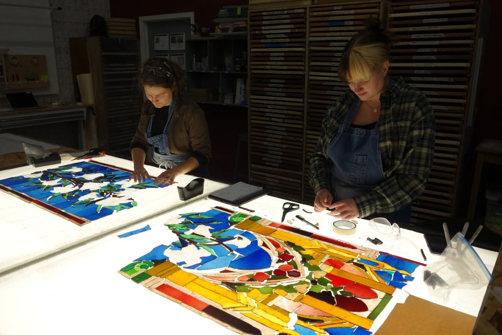 Two artisans working on stained glass restoration, surrounded by colorful glass pieces and tools, in a well-lit workshop dedicated to preserving antique stained glass windows.