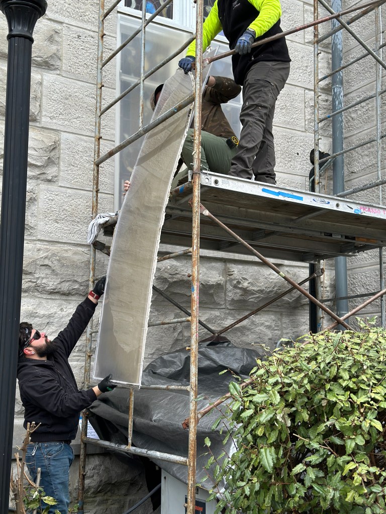 Workers on scaffolding handling protective coverings for stained glass windows, demonstrating maintenance techniques essential for preservation.