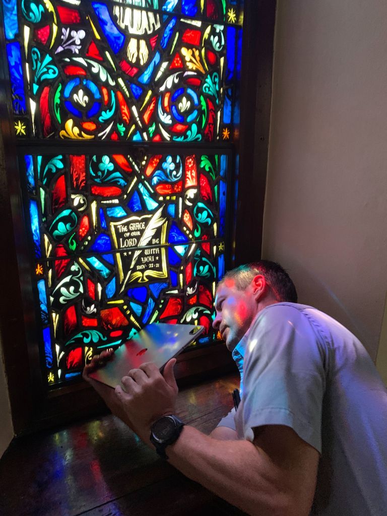 Technician inspecting stained glass window with vibrant colors and religious text, highlighting maintenance efforts for preservation.