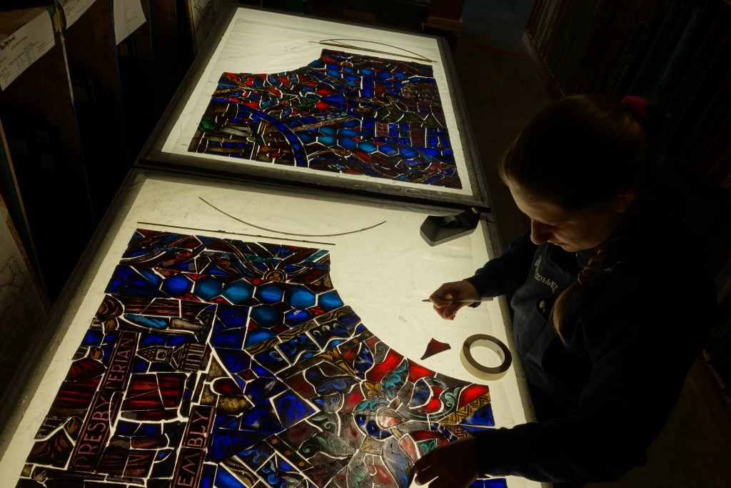 Woman repairing antique stained glass windows, focusing on intricate details and using tools on a workbench, emphasizing preservation efforts for church art.
