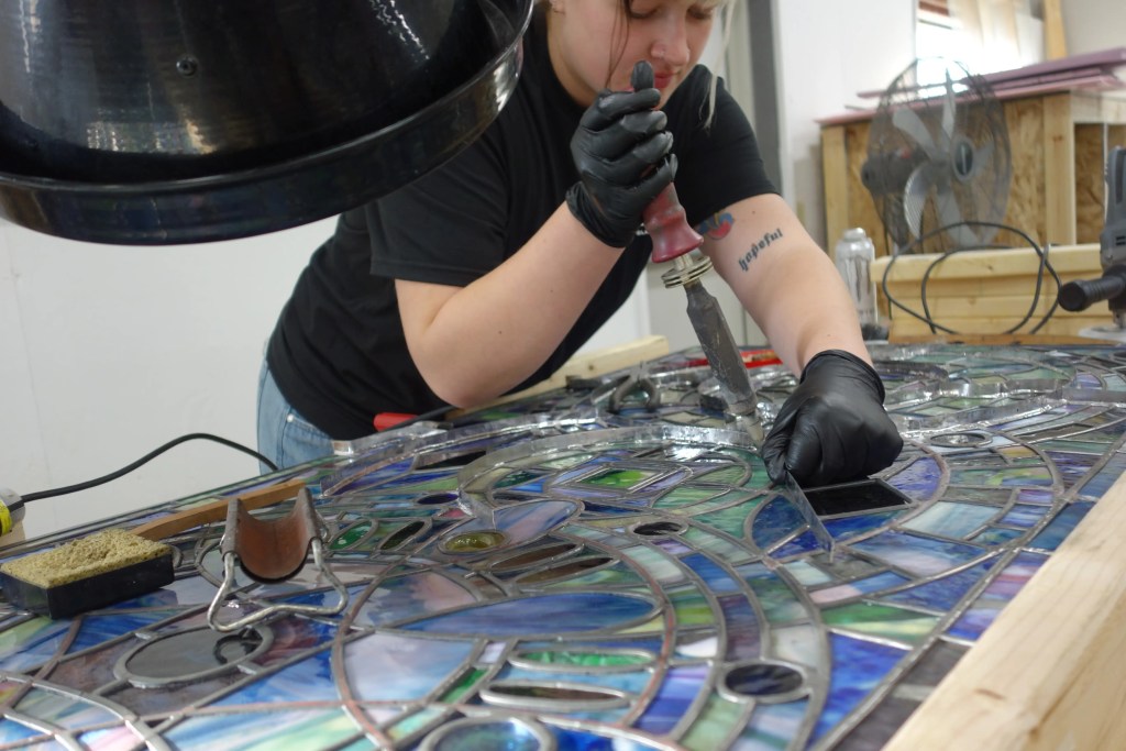 Restoration technician working on stained glass window, applying careful techniques with tools, showcasing intricate colors and patterns in the glass.