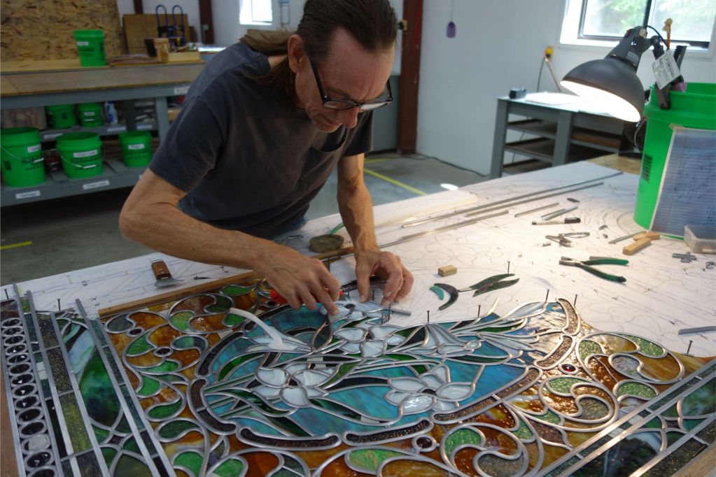 Artisan repairing stained glass window, showcasing detailed craftsmanship and tools, surrounded by vibrant colored glass pieces in a studio setting, reflecting the dedication of Willet Stained Glass Studios to restoration and preservation of sacred art.