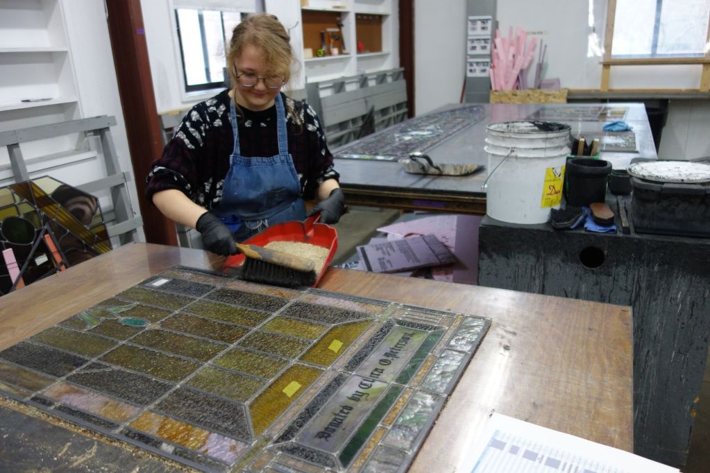 Woman working on stained glass art piece in studio, applying materials to enhance energy efficiency and sustainability in architectural design.