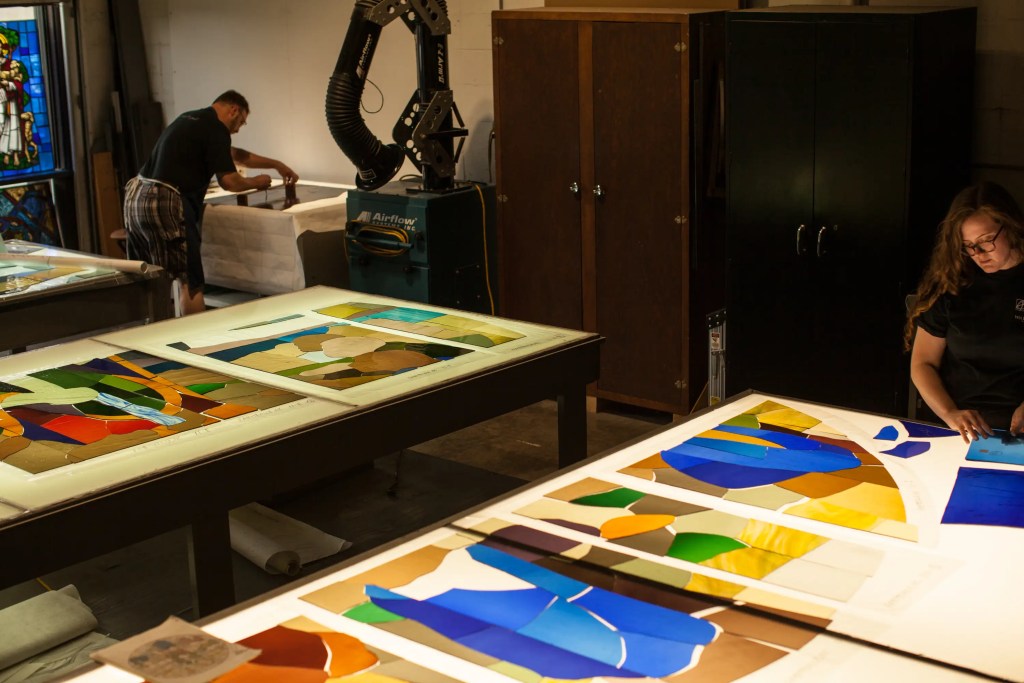 Artisans working on stained glass designs at a light table, showcasing colorful glass pieces and tools in a studio setting, emphasizing craftsmanship and artistry in cathedral glass creation.