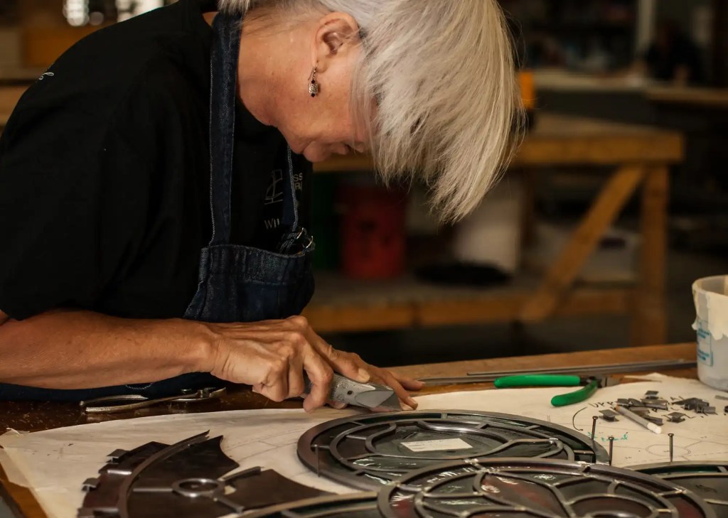 Craftsperson working on stained glass restoration, using tools on a detailed glass panel layout, demonstrating meticulous artistry and technical skill in preservation.
