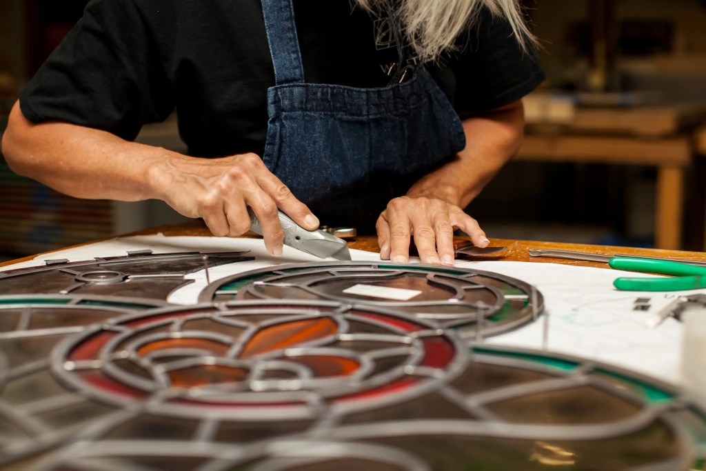 Craftsperson restoring stained glass window, using tools on intricate design, showcasing artistry and technical skill in stained glass restoration.