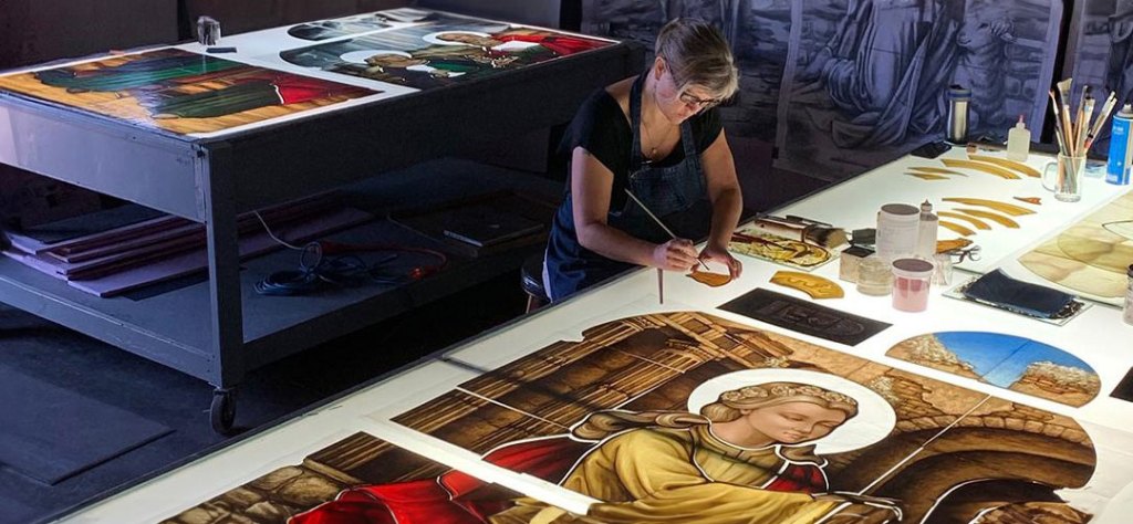Artist restoring stained glass panels, showcasing intricate designs and vibrant colors, with tools and materials for church restoration on a work table.