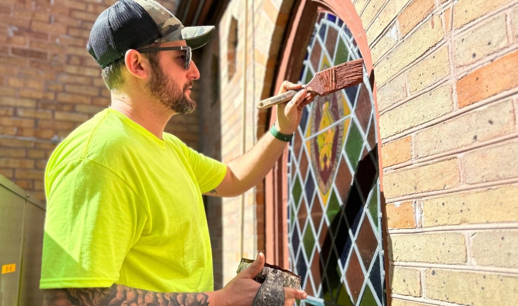 Man in a bright yellow shirt painting a church stained glass window, showcasing restoration efforts for historic glass artwork.