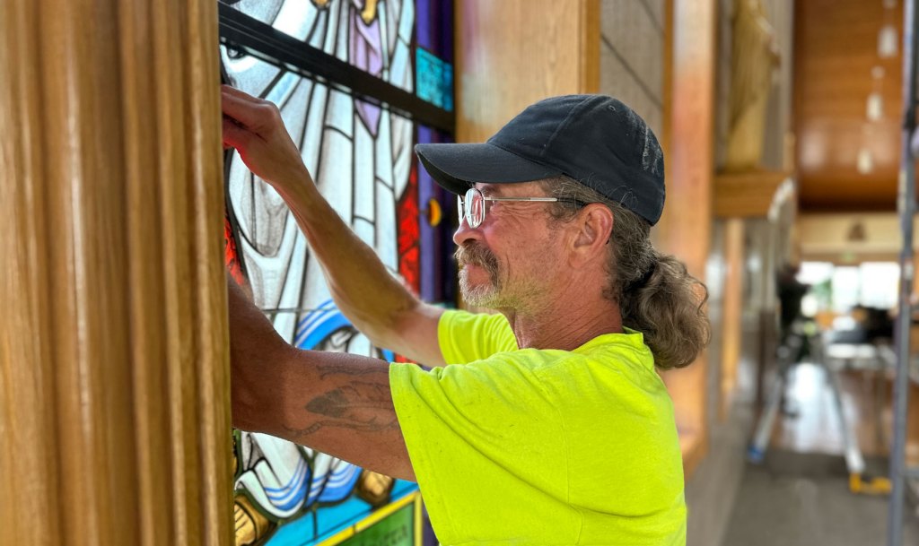 Man installing stained glass window in church, showcasing restoration work on colorful pane, emphasizing craftsmanship and heritage preservation.