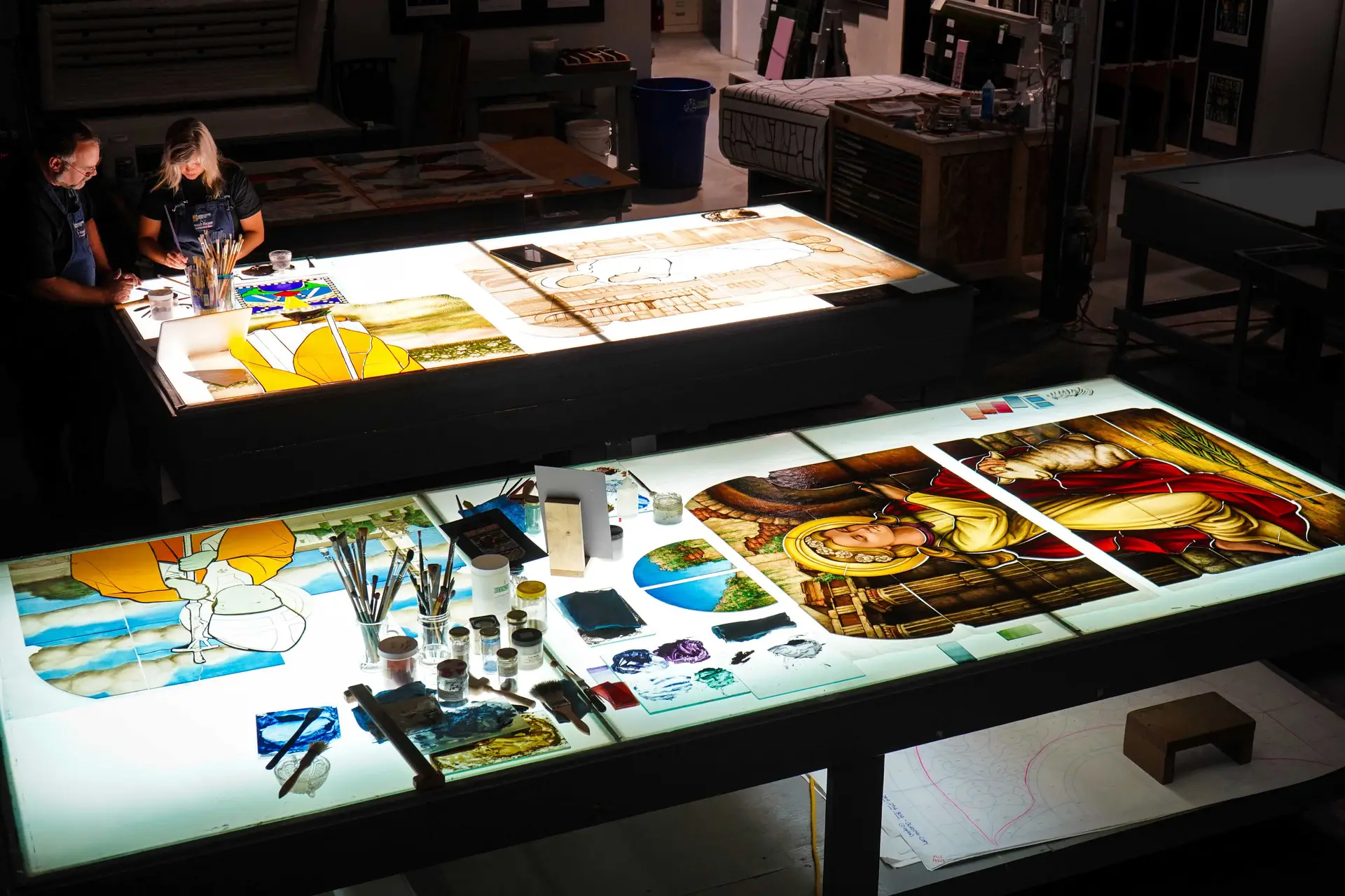 Artists working on custom stained glass designs at illuminated worktables, featuring colorful glass pieces, paintbrushes, and sketches, highlighting the craftsmanship involved in stained glass commissioning.