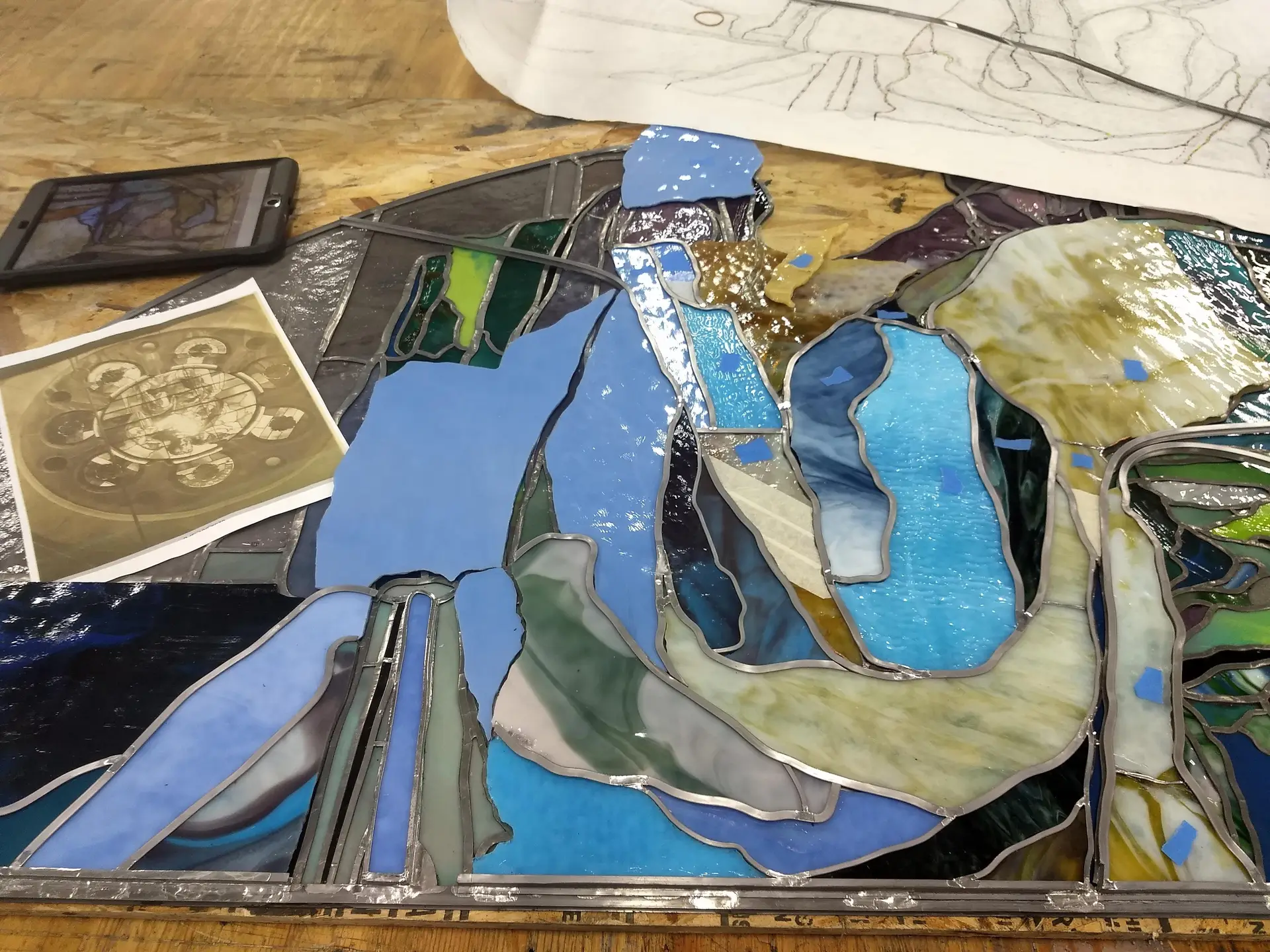 Stained glass panel in progress, featuring vibrant blue and green pieces, with surrounding tools and reference images for restoration techniques.