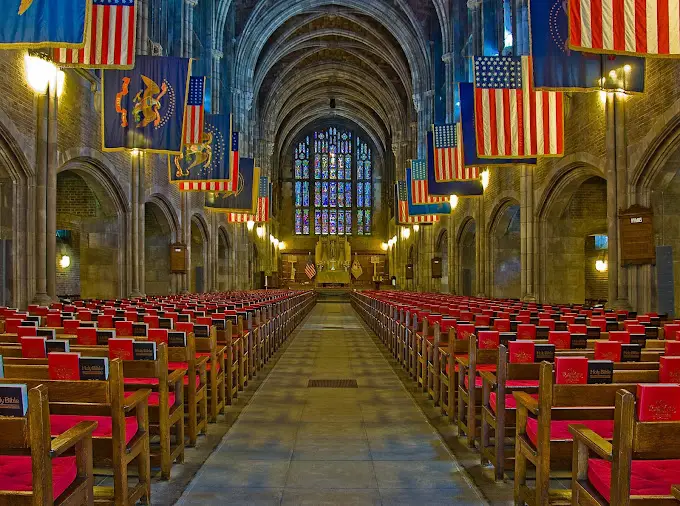 Interior view of a church featuring rows of red cushioned chairs, stained glass windows, and hanging flags, highlighting the importance of stained glass heritage and inspection services.