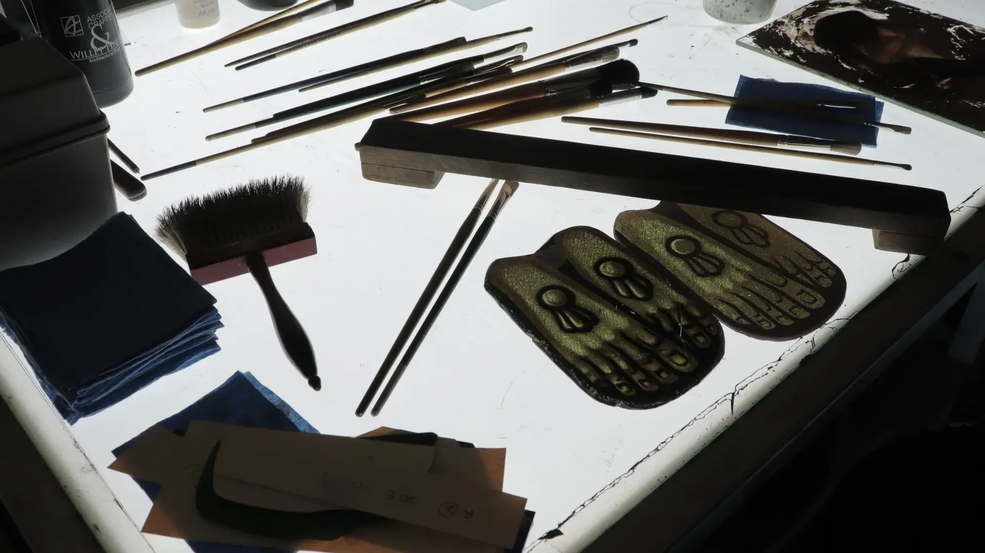 Stained glass conservation tools including brushes, cleaning cloths, and paint palettes on a backlit work surface, highlighting the intricate details of restoration techniques.