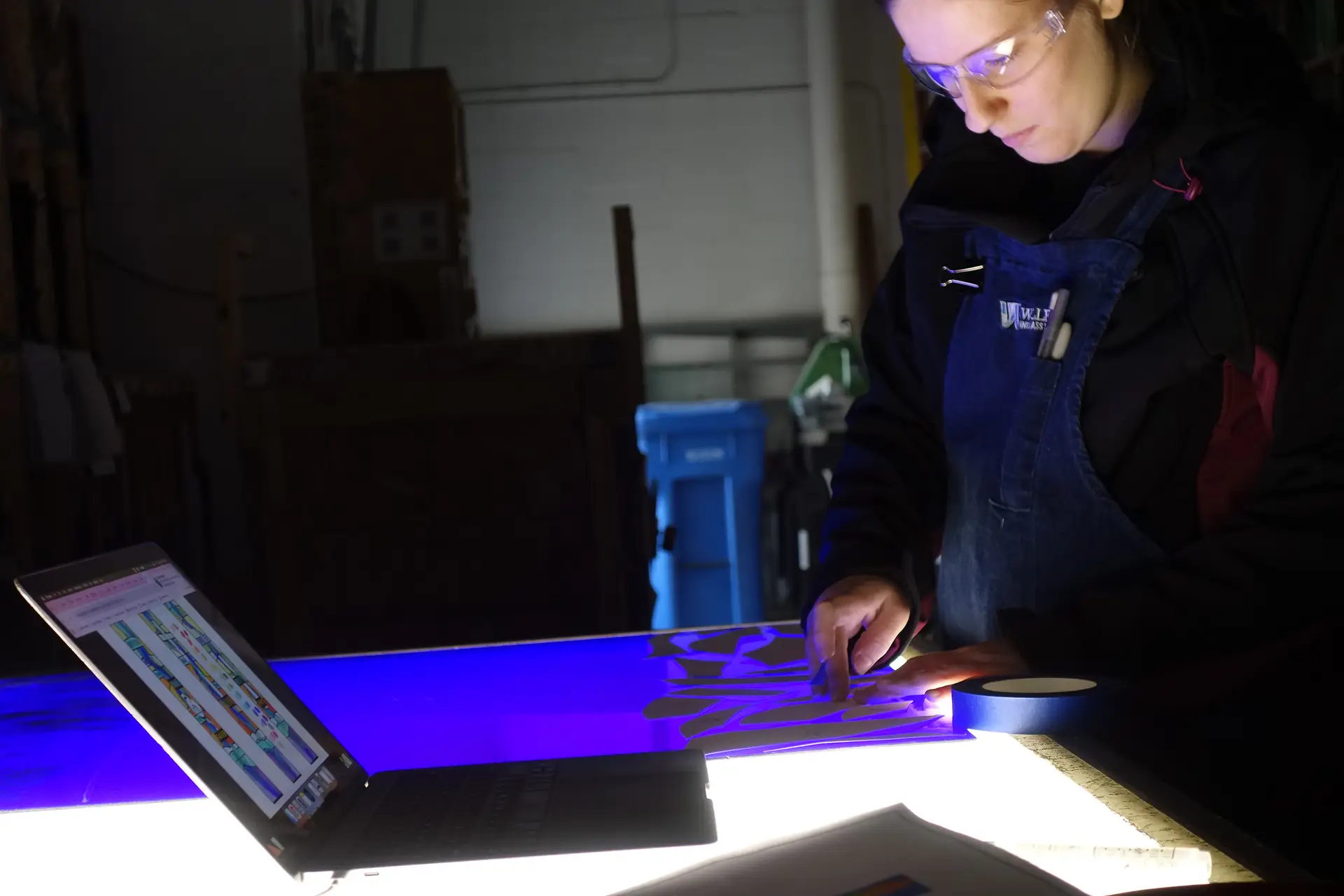 Woman working on stained glass restoration, using tools and materials, with a laptop displaying design plans, illuminated workspace highlighting custom fabrication process.