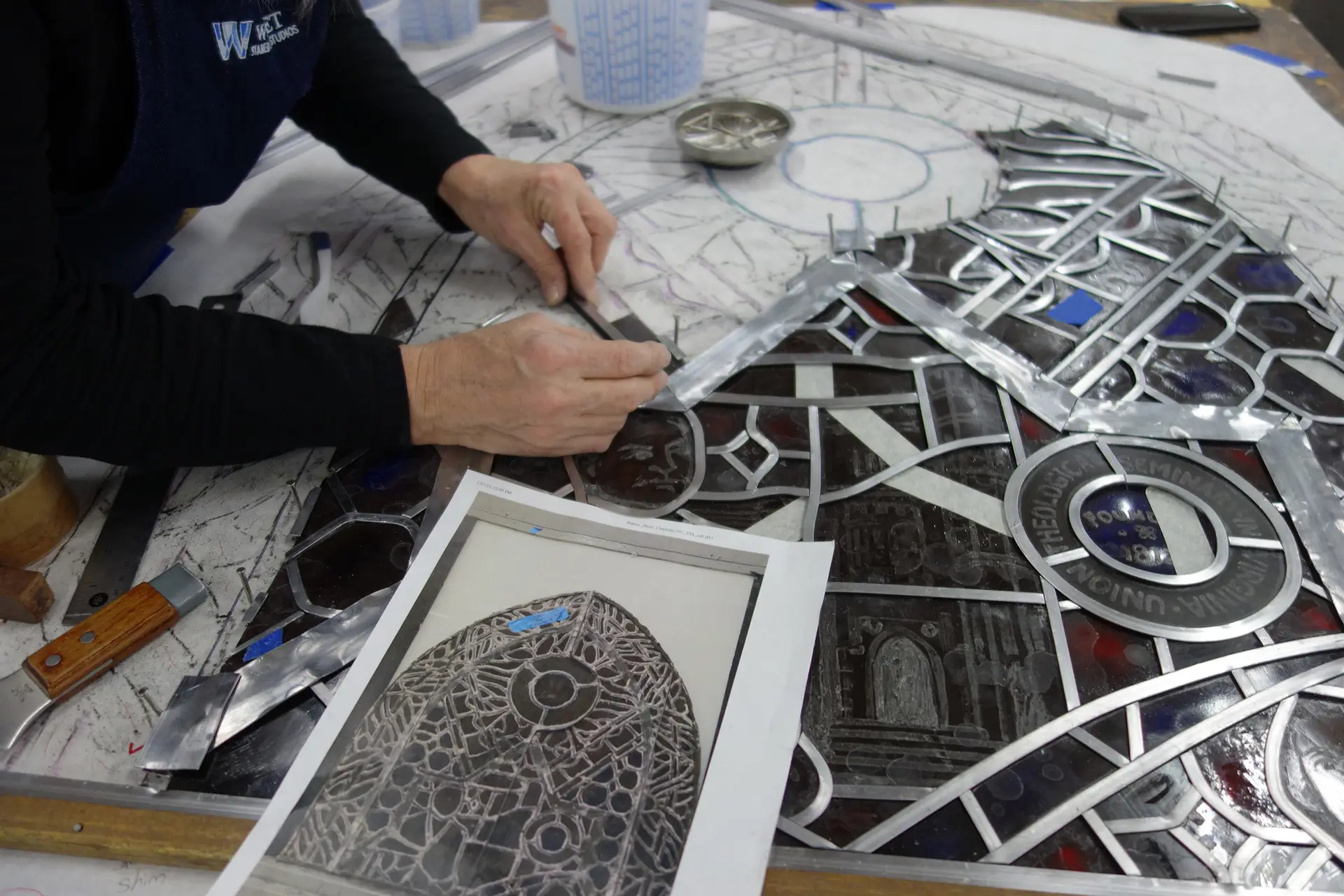 Hands working on stained glass restoration, applying lead came and referencing design plans, showcasing techniques for re-leading and repair.