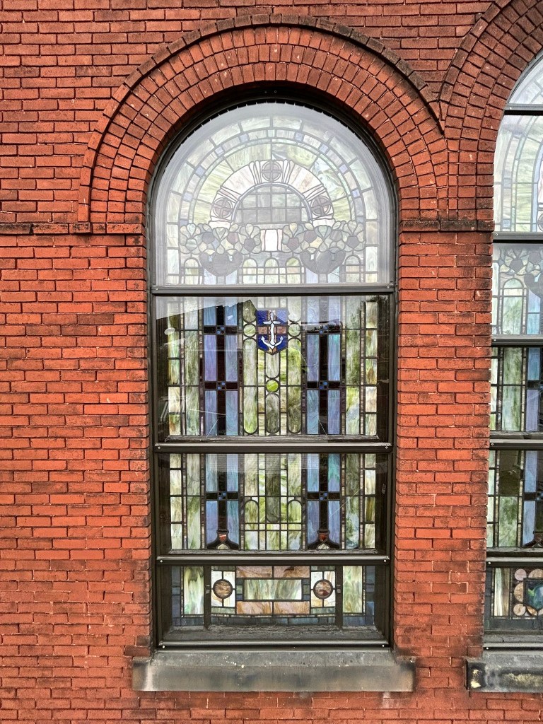 Stained glass window featuring intricate designs and a central emblem, framed by red brick, showcasing restoration work by Willet Studios for Holy Faith Tabernacle Church.