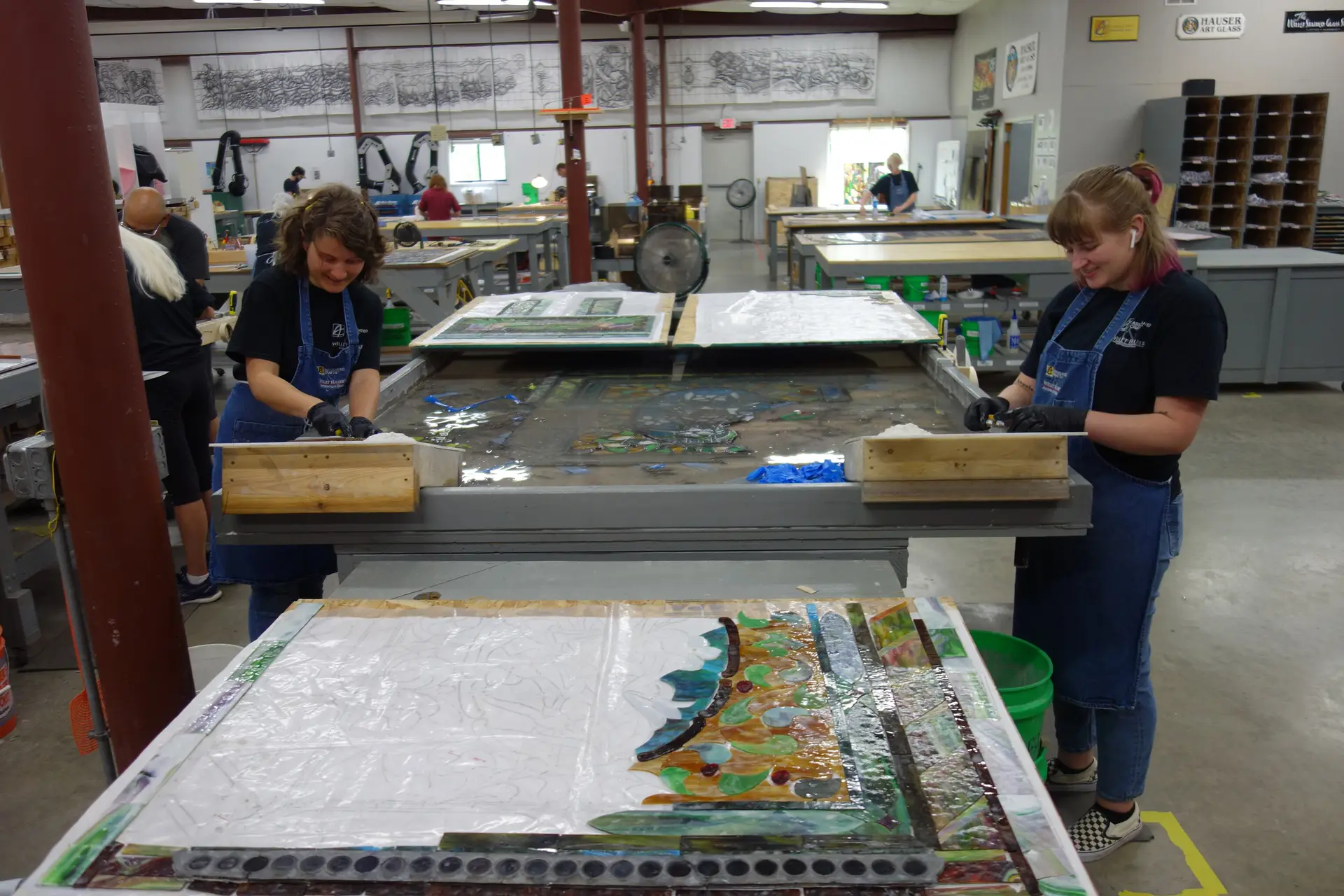 Two professionals working on stained glass restoration in a workshop, using tools to clean and stabilize glass panels, surrounded by various stained glass artworks and conservation equipment.