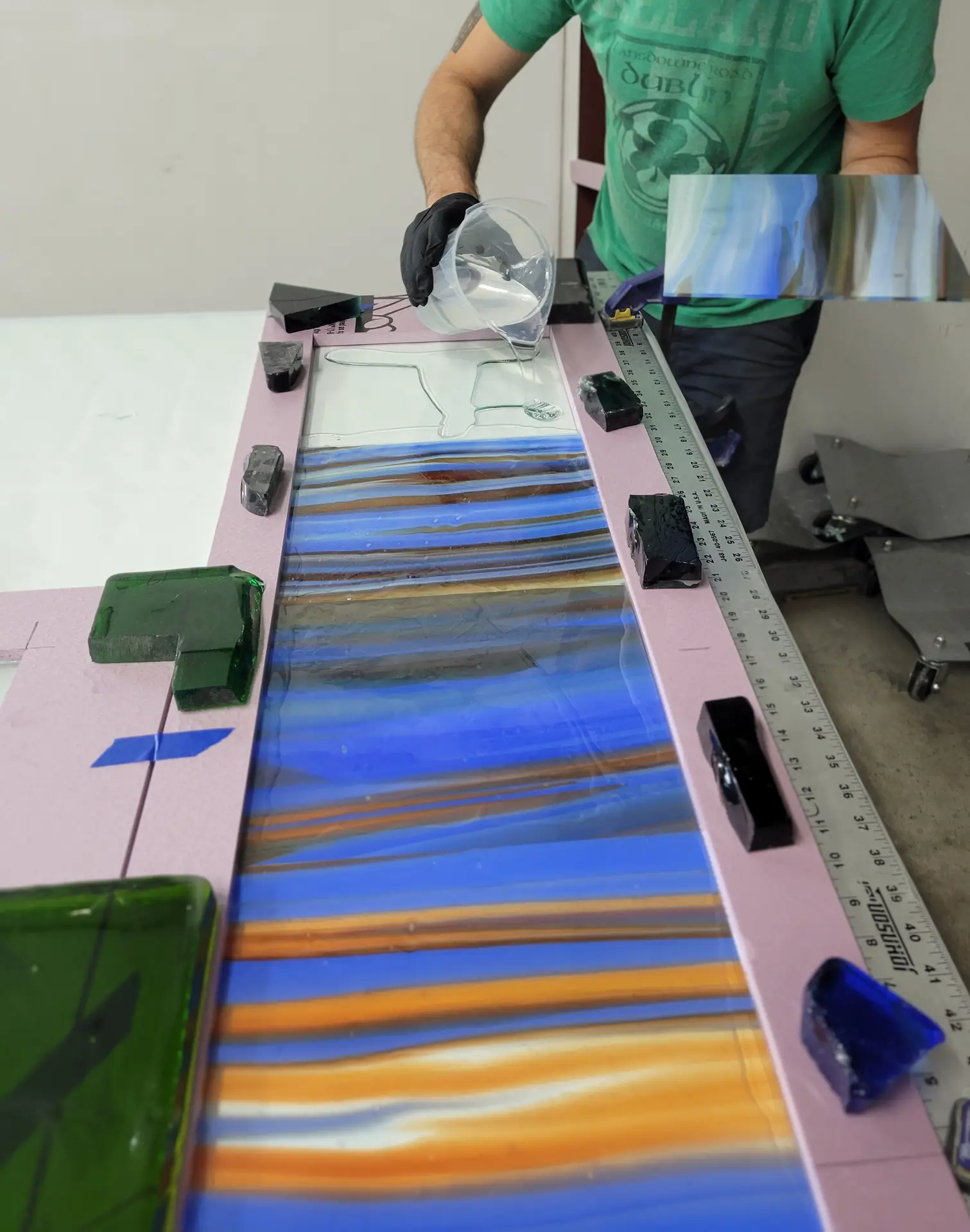Artisan pouring resin onto a custom stained glass panel with vibrant blue and orange patterns, surrounded by green and black glass pieces, showcasing the intricate fabrication process at Willet Studios.