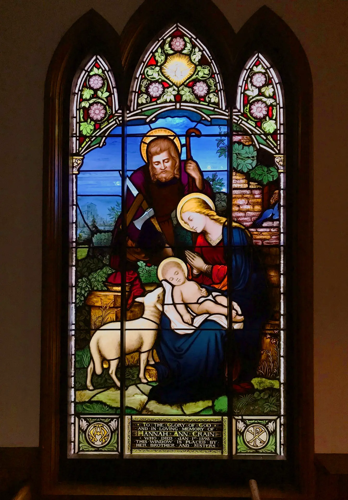 Stained glass window depicting a nativity scene with figures of Mary, Joseph, and baby Jesus, surrounded by a lamb and lush greenery, showcasing intricate craftsmanship and vibrant colors.