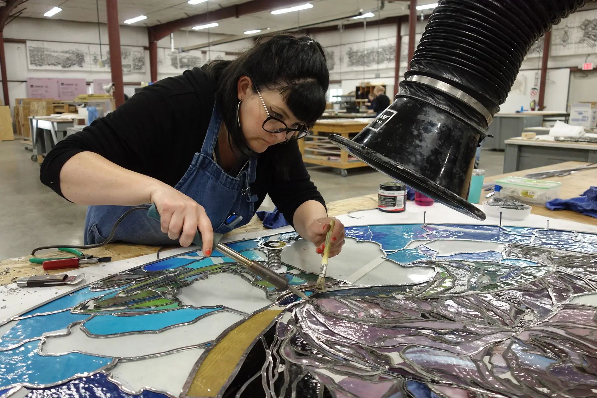 Woman soldering stained glass panel in studio, demonstrating lead came repair techniques, with tools and materials visible, emphasizing stained glass restoration process.