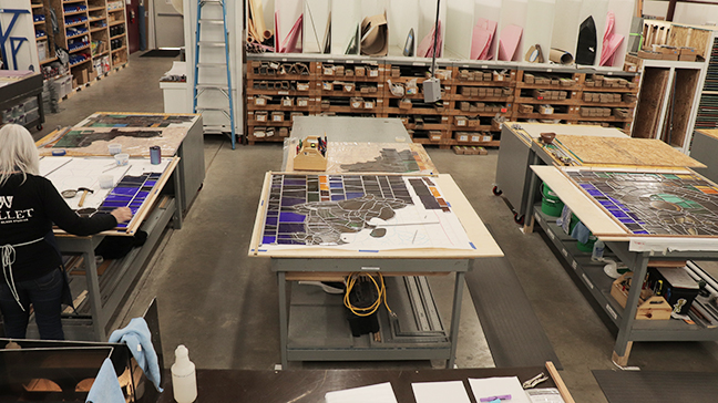 Stained glass workshop with multiple tables displaying glass pieces and tools, artisans working on stained glass designs, emphasizing custom fabrication techniques for ecclesiastical art.