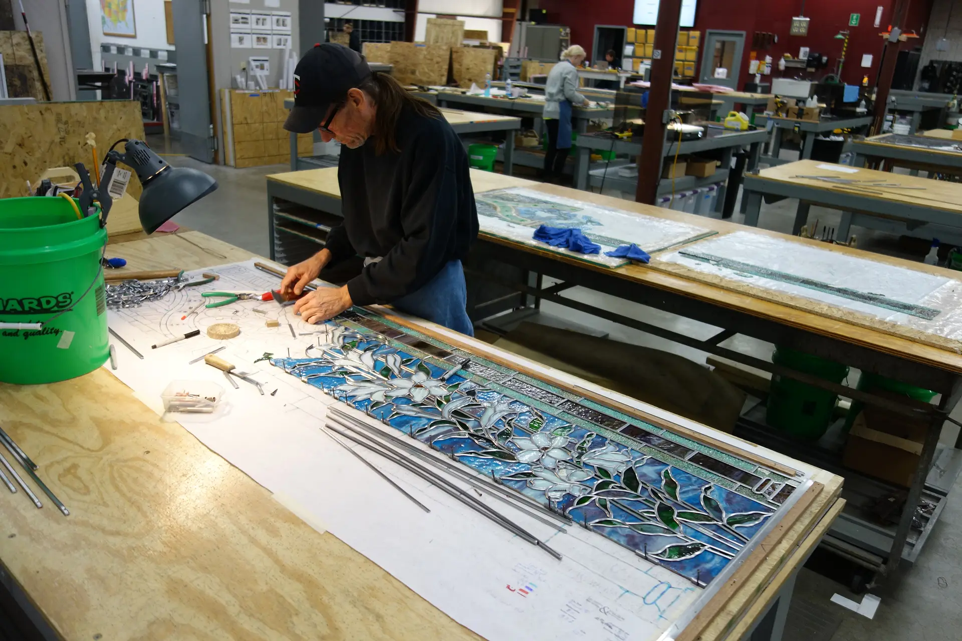 Technician working on stained glass panel repair with tools and materials on a workbench in a stained glass studio.