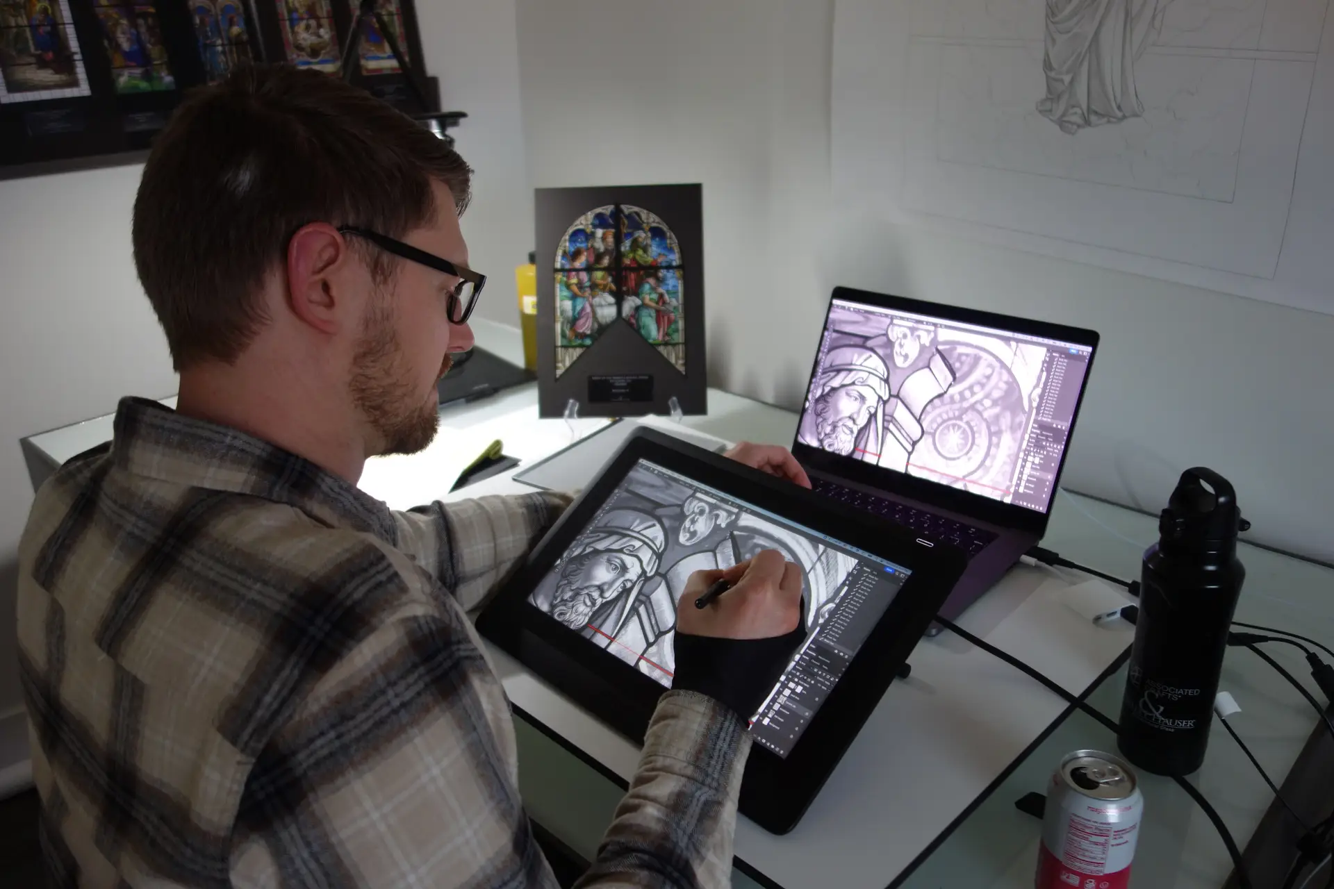 Artist using a digital drawing tablet to design stained glass artwork, with a laptop displaying detailed imagery and a framed stained glass panel in the background, illustrating modern techniques in stained glass design and restoration.