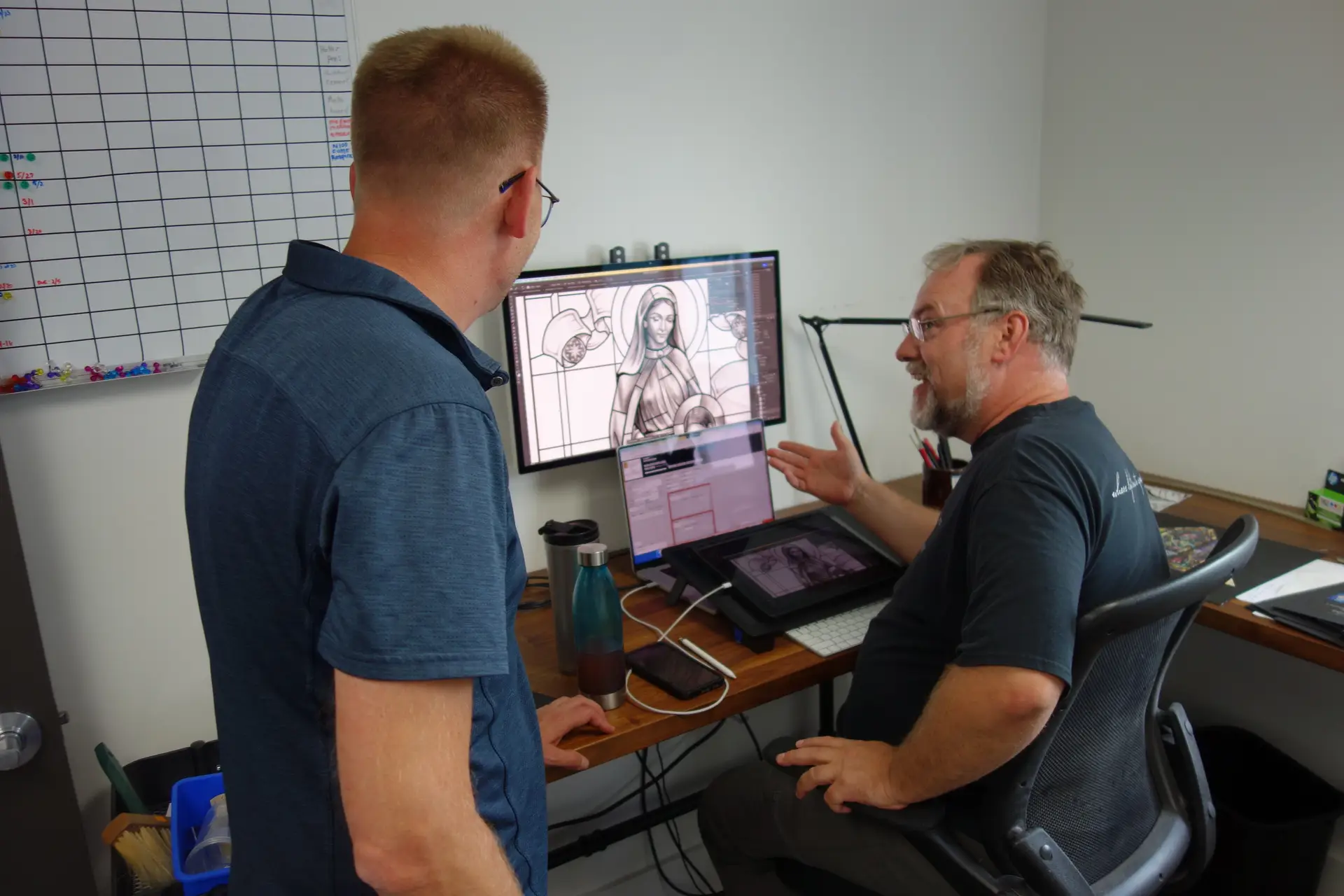 Two men discussing stained glass design on computer screens, one pointing at a design, in a consultation setting at Willet Stained Glass Studios.