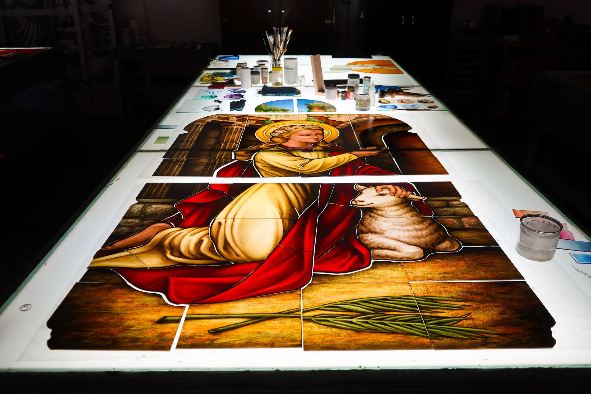 Stained glass panel featuring a figure with a halo, seated beside a lamb, surrounded by tools and materials for stained glass fabrication and restoration.