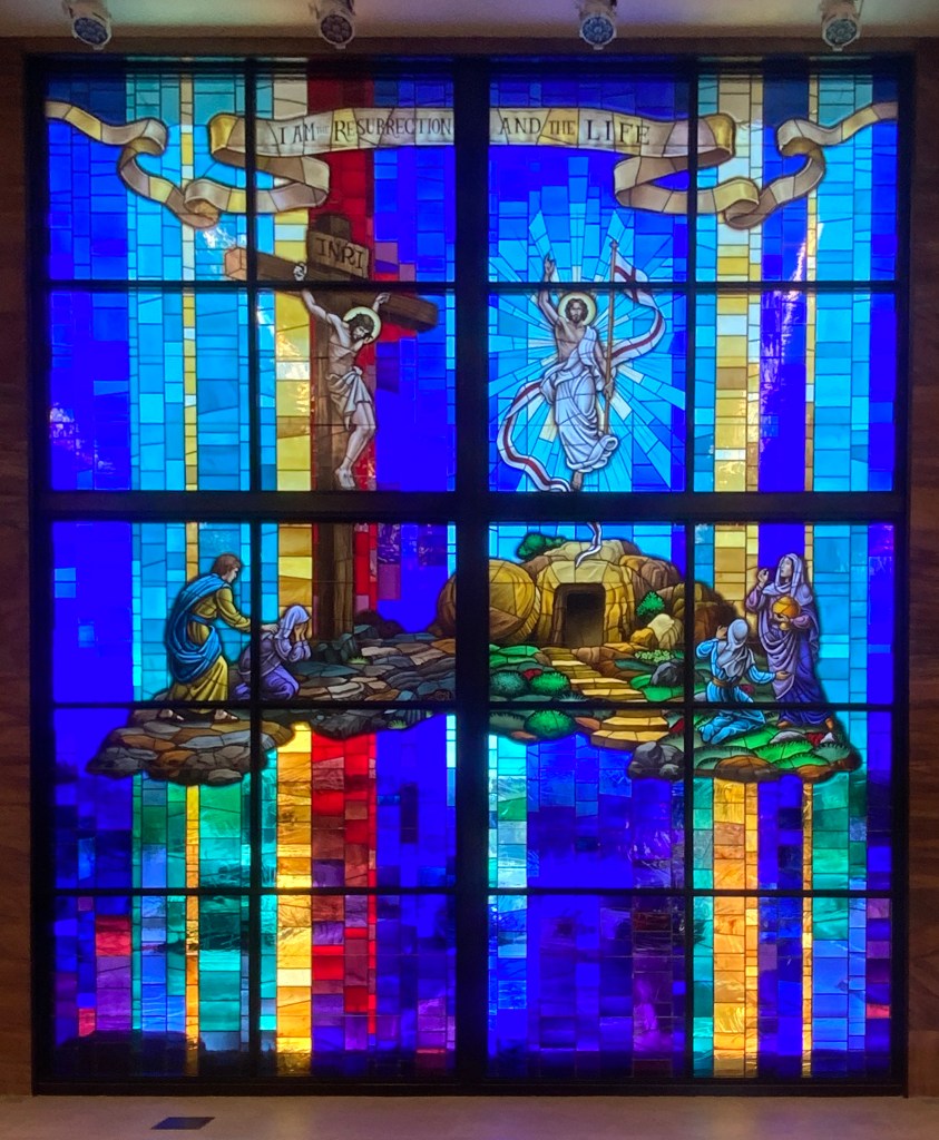 Stained glass window depicting the resurrection scene with vibrant blue, red, and yellow hues, featuring figures of Jesus, Mary, and others, illustrating themes of faith and heritage in stained glass artistry.