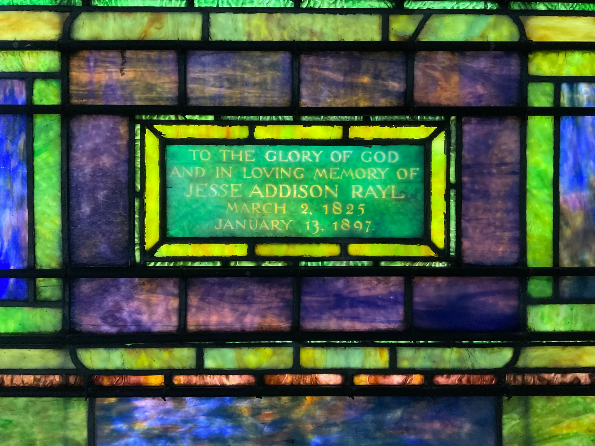 Detailed stained glass window panel with inscription "To the Glory of God and in Loving Memory of Jesse Addison Rayl" and Willet Studios craftsmanship.